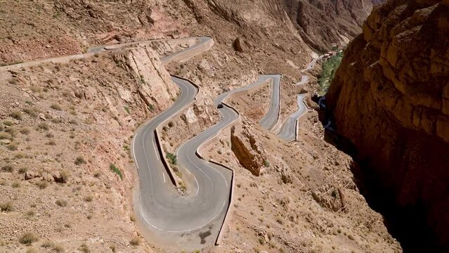 Unidentified car driving through winding road in Dades Gorges, a series of canyons formed by the Dades River. Red and orange rock formations create a dramatic and picturesque landscape.