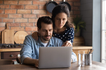 Serious young couple using laptop in cozy kitchen at home, focused woman and man looking at...