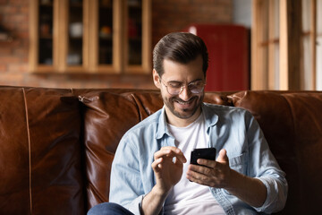 Close up smiling man in glasses having fun with smartphone, sitting on cozy couch at home, spending...