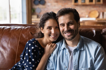 Head shot portrait of happy family hugging, showing love and care, sitting on cozy couch at home together, excited young couple, wife and husband looking at camera, relaxing, enjoying weekends - obrazy, fototapety, plakaty