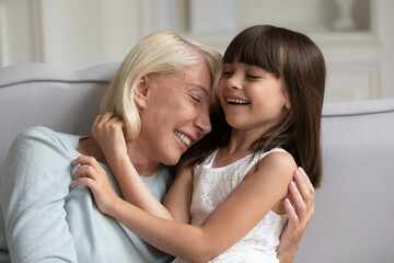 Happy mature grandmother and little granddaughter having fun, sitting on cozy sofa at home, adorable girl grandchild and smiling senior granny cuddling and laughing, spending leisure time together