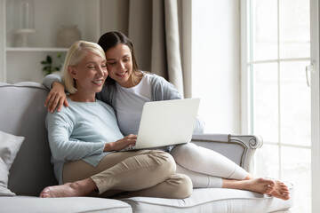 Happy mature mother and adult daughter sitting on couch, using laptop together, smiling grandmother and granddaughter relaxing on cozy sofa, watching video, shopping online, spending weekend at home