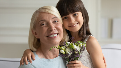 Head shot portrait close up cute little granddaughter presenting flowers to happy grandmother,...