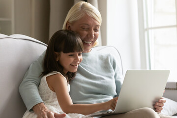 Fototapeta na wymiar Happy mature grandmother and cute little granddaughter having fun with laptop together, looking at screen, sitting on sofa at home, smiling girl grandchild and senior granny using computer close up