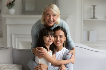 Fototapeta na wymiar Head shot smiling three generations of women at home, happy older grandmother hugging granddaughter and daughter, standing behind cozy sofa, young mother embracing girl child, family portrait