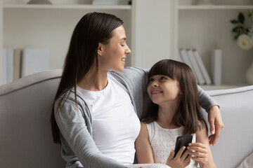 Fototapeta premium Adorable little girl holding mobile phone, sitting on cozy couch with young mother, having fun at home, beautiful mum and cute preschool daughter talking and laughing, spending time together