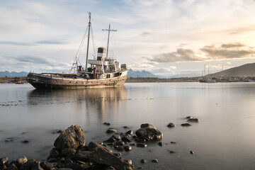 Rocks and a completely calm sea in the foreground with a ship aground with mountains in the...