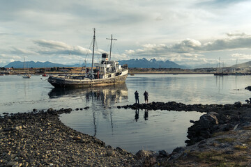 Two people standing on red boats photographing a stranded ship with mountains in the background,...