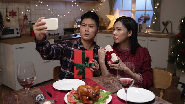 playful japanese couple posing with funny faces and presents while having fun taking selfies at dinner table in the apartment to celebrate Christmas and new year