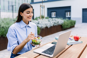 Happy woman holding fresh salad while working on laptop sitting outside building office, looking at screen, watching video or listening colleague on video call. Business concept.