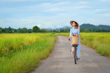 Beautiful Vietnamese Asian woman wearing a blue color Ao Dai National Costume Dress with red bicycle and flowers fresh yellow rice fields mountain background. Portrait fashion show in nature.