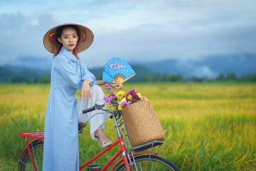 Beautiful Vietnamese Asian woman wearing a blue color Ao Dai National Costume Dress with red bicycle and flowers fresh yellow rice fields mountain background. Portrait fashion show in nature. - obrazy, fototapety, plakaty