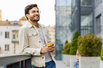 Happy young Indian man enjoying morning coffee hot drink and smiling outdoors. Relaxing, taking a break. Arabian Hindu guy in urban city downtown street, drinking coffee to go. Town lifestyles outside