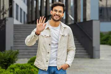 Hello. Indian man smiling friendly at camera, waving hands gesturing hi, greeting or goodbye,...