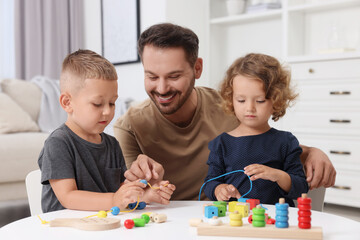 Motor skills development. Father and his kids playing with wooden pieces and string for threading activity at table indoors