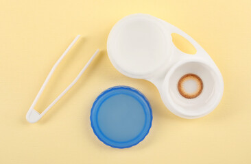 Case with color contact lenses and tweezers on pale yellow background, flat lay