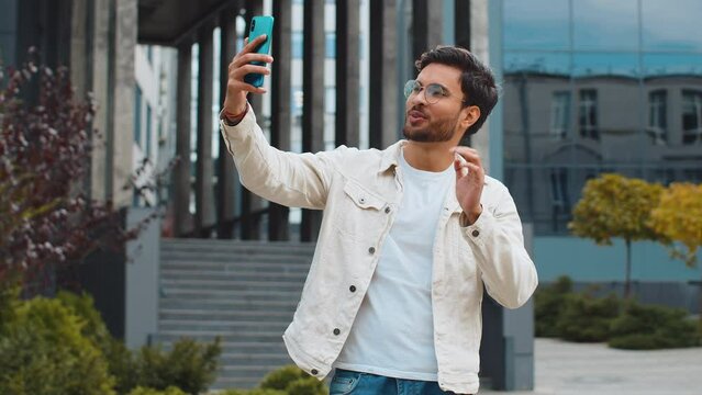 Young Indian man blogger taking selfie on smartphone, communicating video call online with subscribers recording stories for social media vlog outdoors. Arabian Hindu guy standing in urban city street