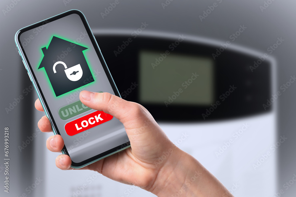 Poster woman operating home alarm system via mobile phone against grey wall with security control panel, cl