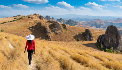 A woman with a red long-sleeved shirt and a white hat walks on golden dry grasslands at the ridge of Mulayit Taung, Myanmar. There are overlapping hills and many rocks. There is copy space on top