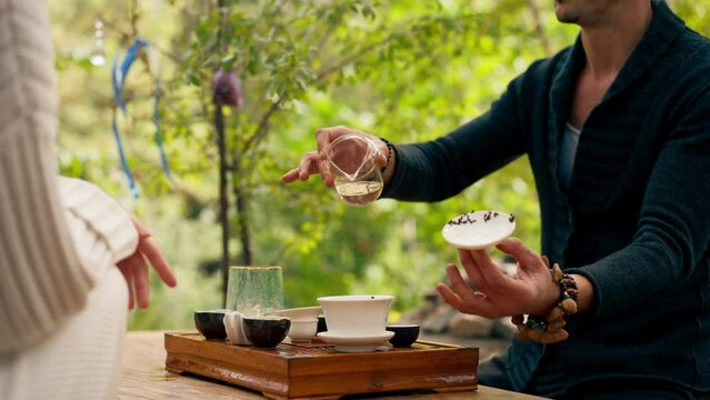 close-up The hands of a professional tea master who pours fresh natural green tea from a glass teapot into bowls on wooden chaban table