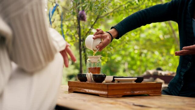 close-up The hands of a professional tea master who pours fresh natural green tea from a glass teapot into bowls on wooden chaban table