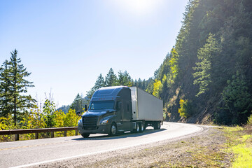 Fototapeta na wymiar Stylish dark gray big rig semi truck transporting cargo in dry van semi trailer running on the winding mountain road