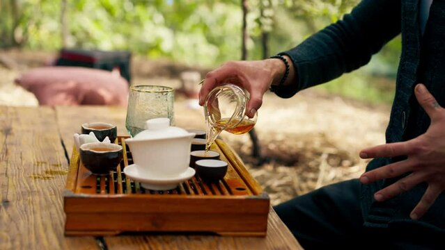 close-up The hands of a professional tea master who pours fresh natural green tea from a glass teapot into bowls on wooden chaban table