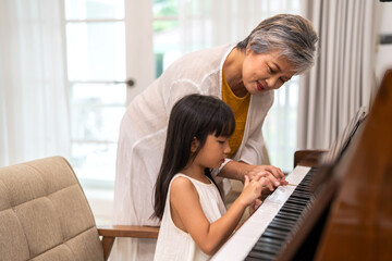 Portrait of happy love asian grandmother and asian cute girl play and enjoy relax teaching and play piano music lesson at home.senior, insurance, care.girl with their laughing grandparents.Family. - obrazy, fototapety, plakaty