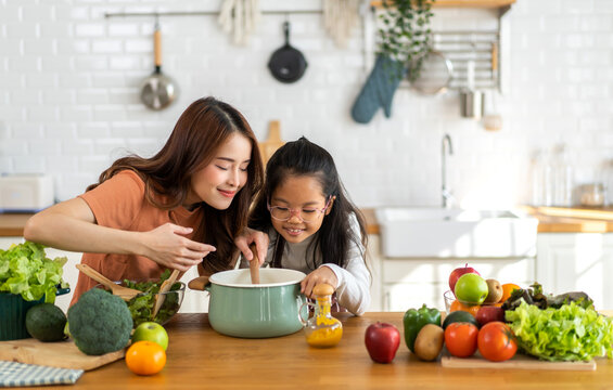 Portrait Of Happy Love Asian Family Mother With Little Asian Girl Daughter Child Help Cooking Food Healthy Eat With Fresh Vegetable Testing Smell Soup In A Pot With Spoon.help Mommy In Kitchen At Home