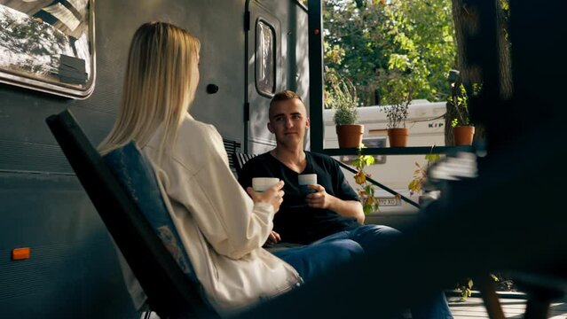 A young handsome guy and a girl are sitting on the veranda of a mobile home in camper drinking tea and talking enthusiastically