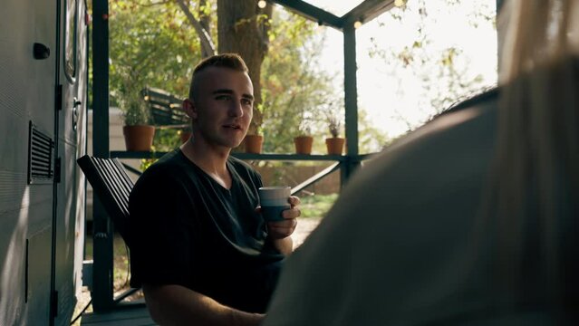 A young handsome guy and a girl are sitting on the veranda of a mobile home in camper drinking tea and talking enthusiastically