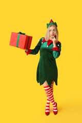 Young woman in elf costume with Christmas gift box on yellow background