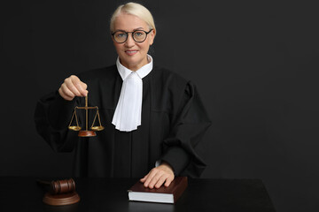 Mature female judge with scales of justice and book at table on black background