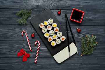 Board of tasty sushi rolls with fir branches and Christmas decorations on black wooden background