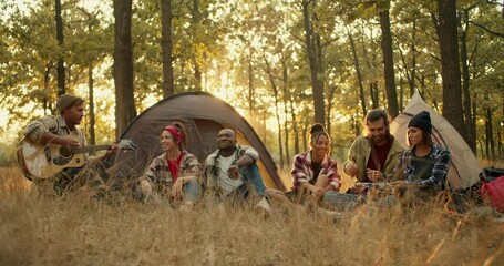 A company of 6 trekking participants in special hiking clothes against the backdrop of tents. A blond man in a hat plays an acoustic guitar and all the other participants in the hike sing along and