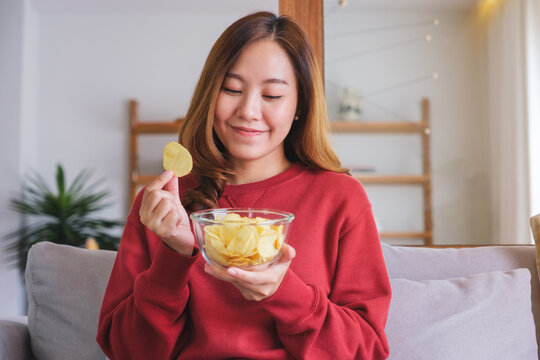 Portrait image of a young woman picking and eating potato chips at home