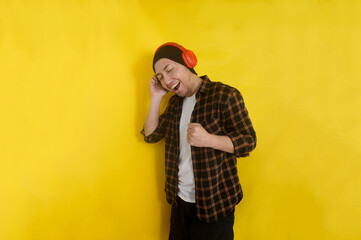Excited young Asian man wearing beanie hat, listening music with headphones and sing isolated over yellow background