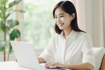 Beautiful asian woman in the white shirt is smiling and working on her laptop at home. Shallow depth of field, selective focus
