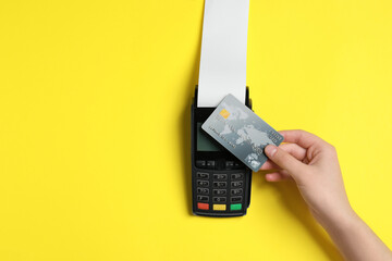 Woman using payment terminal with credit card and thermal paper for receipt on yellow background,...