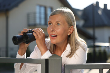 Concept of private life. Curious senior woman with binoculars spying on neighbours over fence outdoors