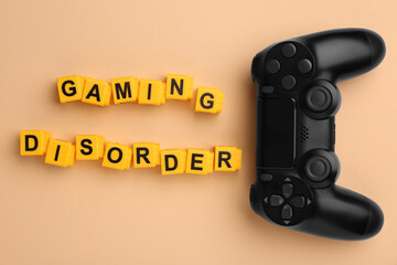 Phrase Gaming Disorder made of yellow cubes and gamepad on beige background, flat lay. Addictive...