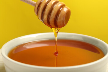 Pouring tasty honey from dipper into bowl against yellow background, closeup