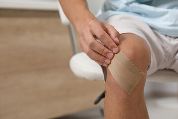 Man putting sticking plaster onto knee indoors, closeup. Space for text