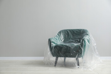 Stylish armchair covered with plastic film near light grey wall indoors. Space for text