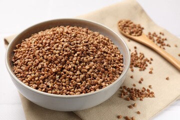 Bowl with dry buckwheat, spoon and fabric on white table, closeup