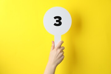 Woman holding auction paddle with number 3 on yellow background, closeup - obrazy, fototapety, plakaty