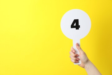 Woman holding auction paddle with number 4 on yellow background, closeup. Space for text