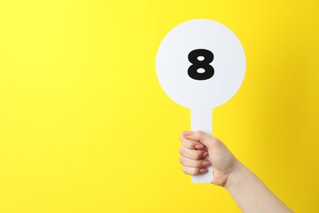 Woman holding auction paddle with number 8 on yellow background, closeup. Space for text