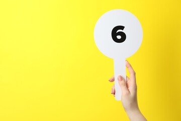 Woman holding auction paddle with number 6 on yellow background, closeup. Space for text