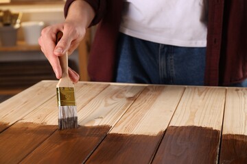 Fototapeta na wymiar Man with brush applying wood stain onto wooden surface indoors, closeup